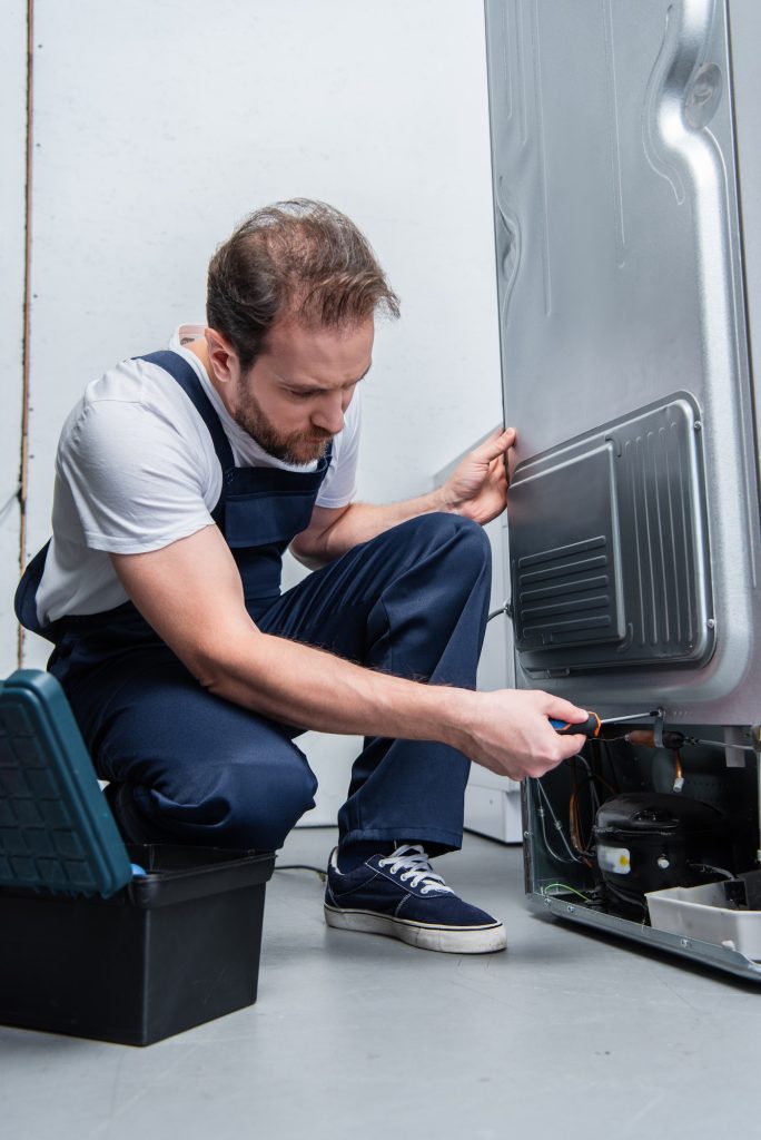 repairman in working overall fixing refrigerator by screwdriver in kitchen